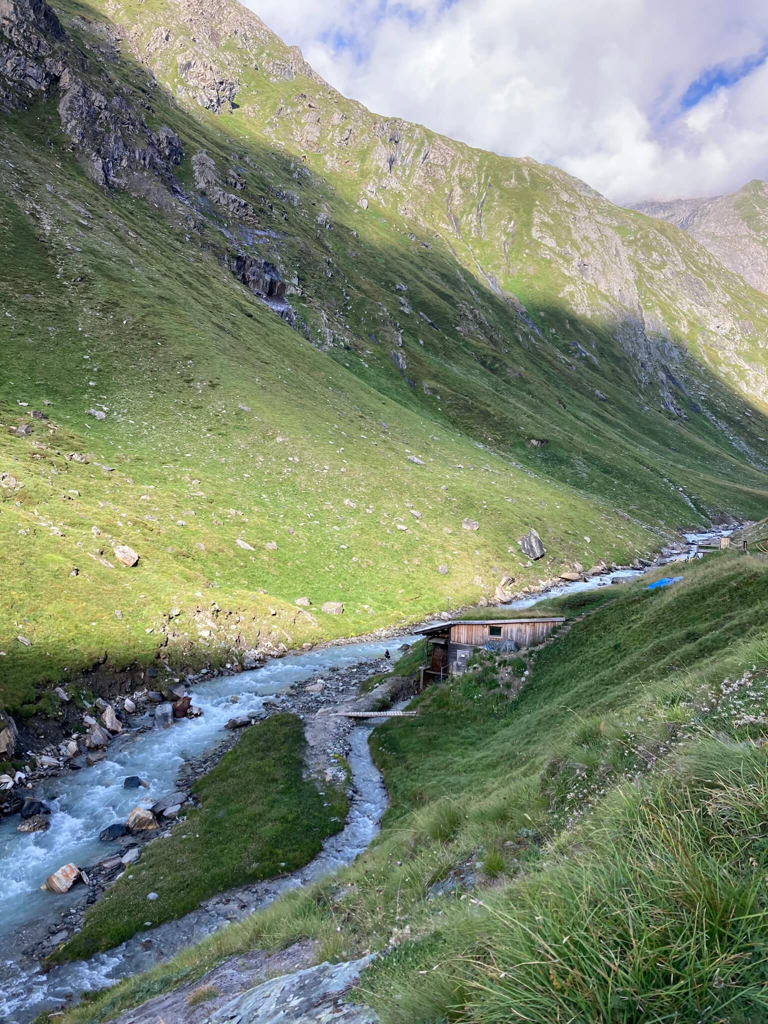 Bild zeigt die Bergwelt und eine Holzhütte am Fluss.